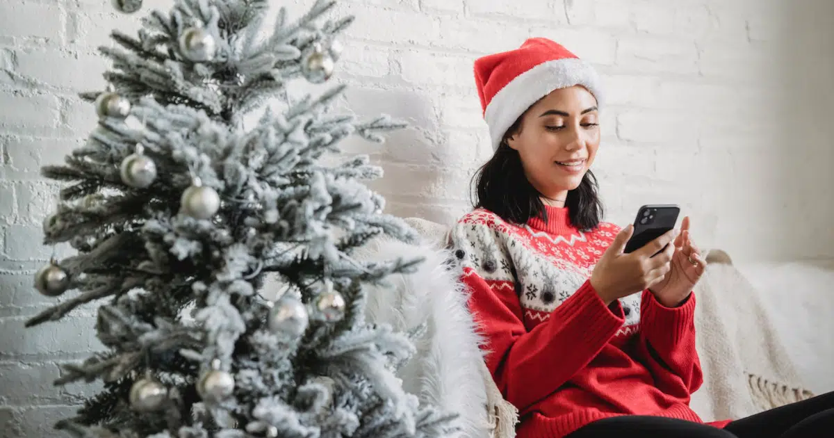 woman with smartphone sitting next to a christmas tree